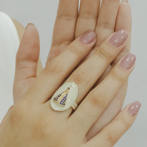 Foto das mãos de uma mulher de pele clara, com unhas em rosa nude,usando o anel de nossa senhora aparecida oval, em resina branca, com manto em zircônia azul. Foto está demonstrando como fica o anel em uso real.