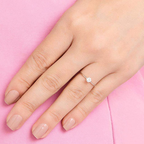Mulher da cor clara com uma roupa rosa tendo foco nas mãos da moça, que está com a unha clara para mostrar um anel prateado com uma pedra de brilhantes nele.