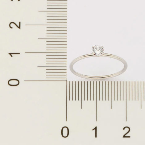 Fundo cinza com uma régua na frente da cor preta, e um pouco em cima tem um anel prateado com uma pedra de brilhantes sendo medido.