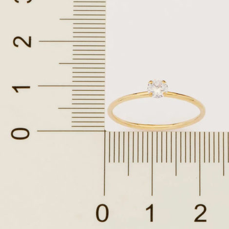 Fundo cinza com uma régua na frente na cor preta, um pouquinho em cima da régua tem um anel da cor dourado com uma pedra de brilhantes branca.