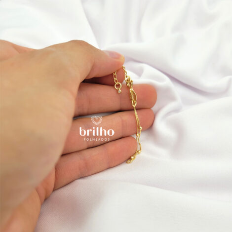 Foto de mão de pele clara segurando a pulseira de bolinhas infantil em cima de tecido sedoso branco, semijoia da marca Sabrina Joias revendida pela loja Brilho Folheados. Código 1700293