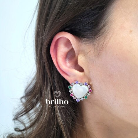 Foto de orelha de modelo de pele clara usando maxi brinco coração em madrepérola com zircônias coloridas da marca Sabrina Joias. Código R1690968