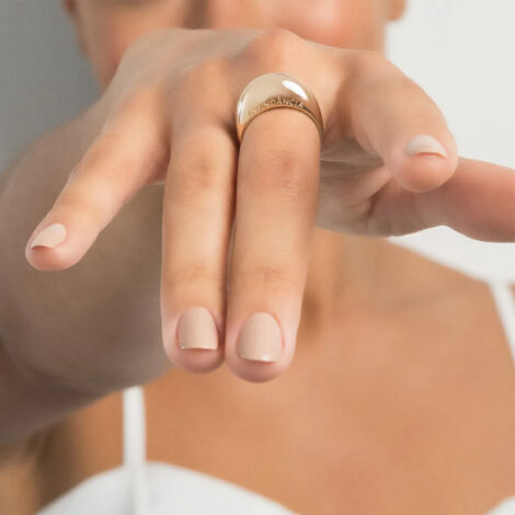 Foto de mão de modelo de pele clara usando o anel abaulado com escrita abundancia folheado a ouro, cor dourado. Código 513318